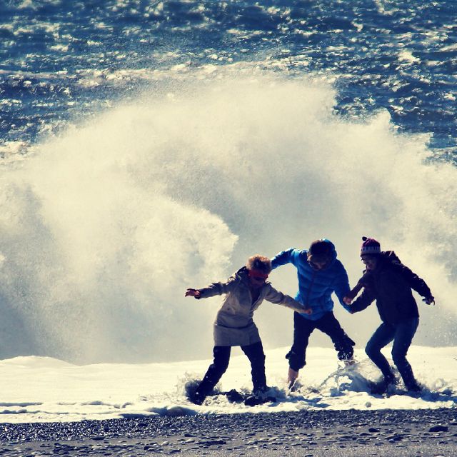 3 Personen am Strand vor einer Welle.