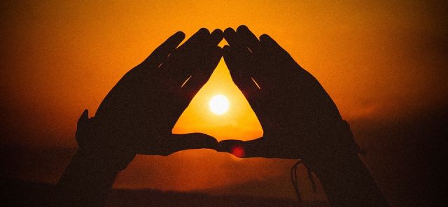 Das Bild zeigt zwei Hände die ein Dreieck formen. In der Mitte sieht man den Sonnenuntergang.
