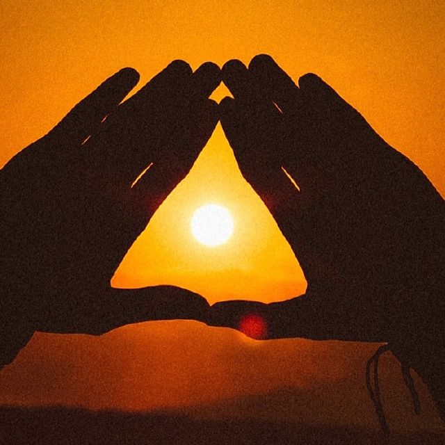 Das Bild zeigt zwei Hände die ein Dreieck formen. In der Mitte sieht man den Sonnenuntergang.