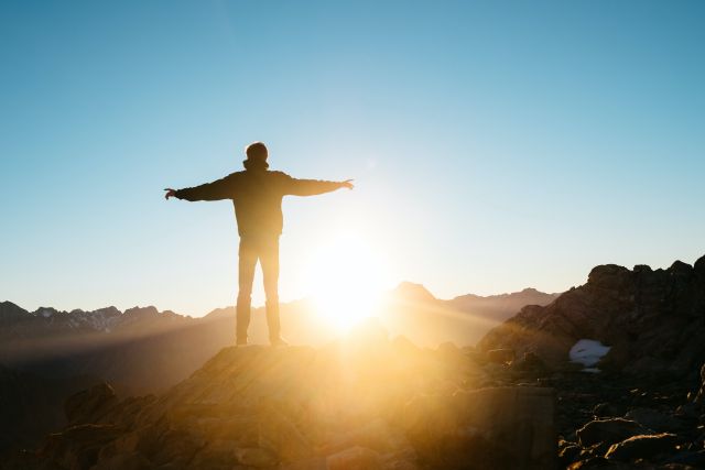 Ein Mann steht auf einem Berg und blickt mit ausgestreckten Armen in den Sonnenaufgang