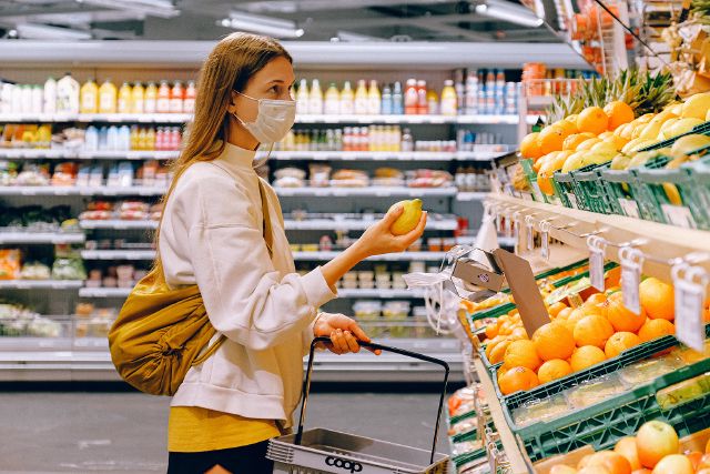 Eine Frau steht mit Maske im Supermarkt und hat eine Zitrone in der Hand.