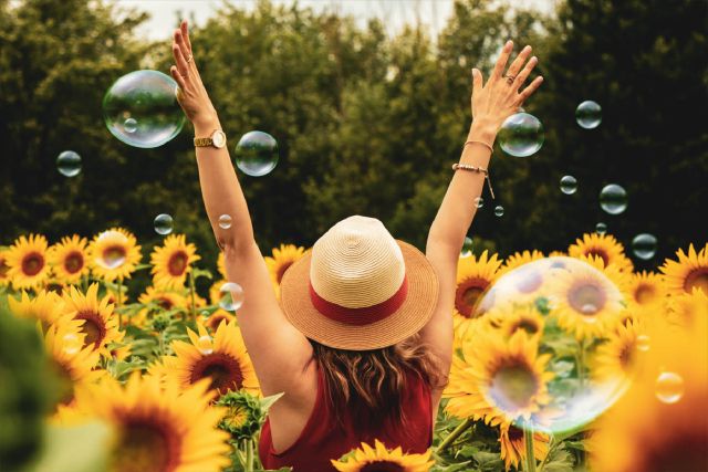 Eine Frau steht in einem Sonnenblumenfeld zwischen Seifenblasen und streckt die Arme in die Luft.